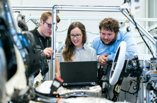 Woman Surrounded by 2 Men Looking at a Laptop Screen and Surrounded by Tech Hardware