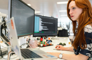 Woman with Red Hair Sitting at a Desk and Looking at 2 Computer Monitors
