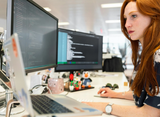 Woman with Red Hair Sitting at a Desk and Looking at 2 Computer Monitors