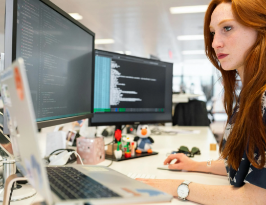 Woman with Red Hair Sitting at a Desk and Looking at 2 Computer Monitors