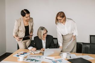 3 Business Women Surrounded By Paperwork and Looking at a Laptop Screen