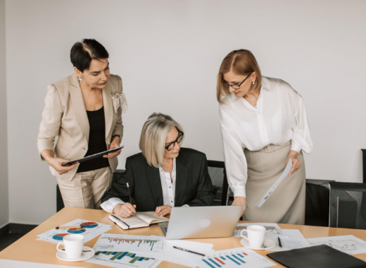 3 Business Women Surrounded By Paperwork and Looking at a Laptop Screen