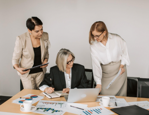 3 Business Women Surrounded By Paperwork and Looking at a Laptop Screen