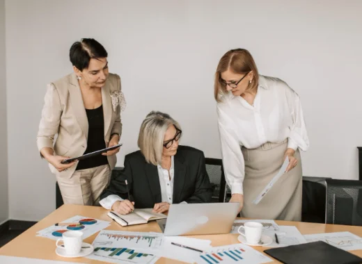 3 Business Women Surrounded By Paperwork and Looking at a Laptop Screen