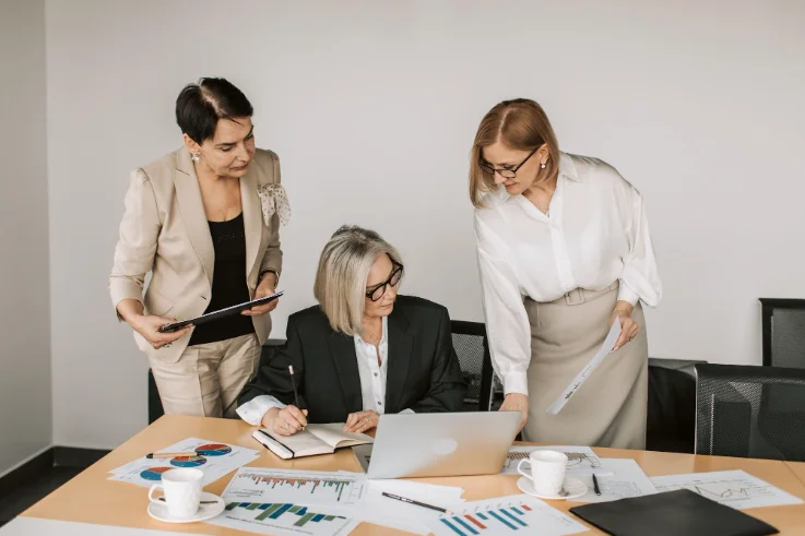 3 Business Women Surrounded By Paperwork and Looking at a Laptop Screen
